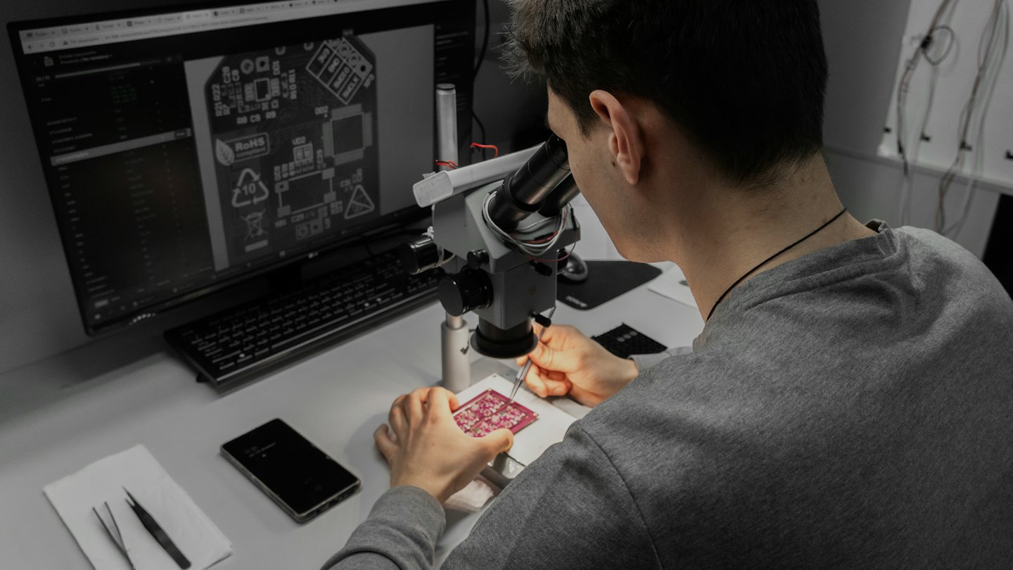 Uomo ispeziona attentamente un circuito stampato al microscopio su una scrivania.
